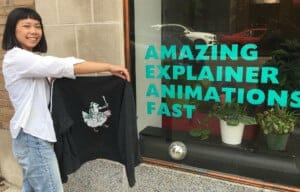 Jacquie poses in front of a glass storefront window that reads, Amazing Explainer Animations Fast; she is holding the Chicken Knight sweatshirt