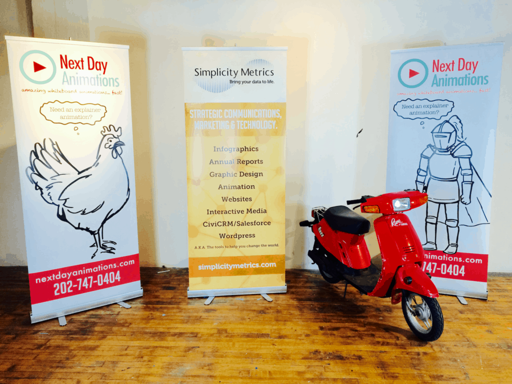 3 poster boards, one featuring a chicken and one featuring a knight both featuring NDA logos. a third reads "Simplicity Metrics." A red scooter sits next to the posters.