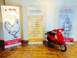 3 poster boards, one featuring a chicken and one featuring a knight both featuring NDA logos. a third reads "Simplicity Metrics." A red scooter sits next to the posters.